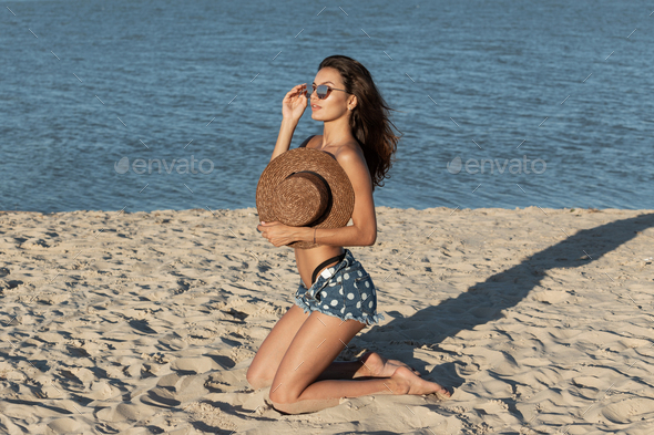 Dark-haired girl in jean shorts kneels in sunglasses on the sand near the sea and hold a hat on her - Stock Photo - Images