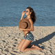 Dark-haired girl in jean shorts kneels in sunglasses on the sand near the sea and hold a hat on her - PhotoDune Item for Sale