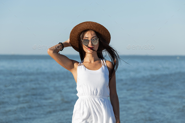 Stunning dark-haired girl in white dress, sunglasses and brown hat near the sea on a sunny day - Stock Photo - Images