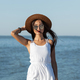 Stunning dark-haired girl in white dress, sunglasses and brown hat near the sea on a sunny day - PhotoDune Item for Sale