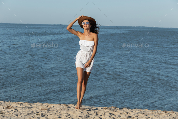 Gorgeous dark-haired girl in white dress, hat and sunglasses walk on the sandy beach near the sea on - Stock Photo - Images