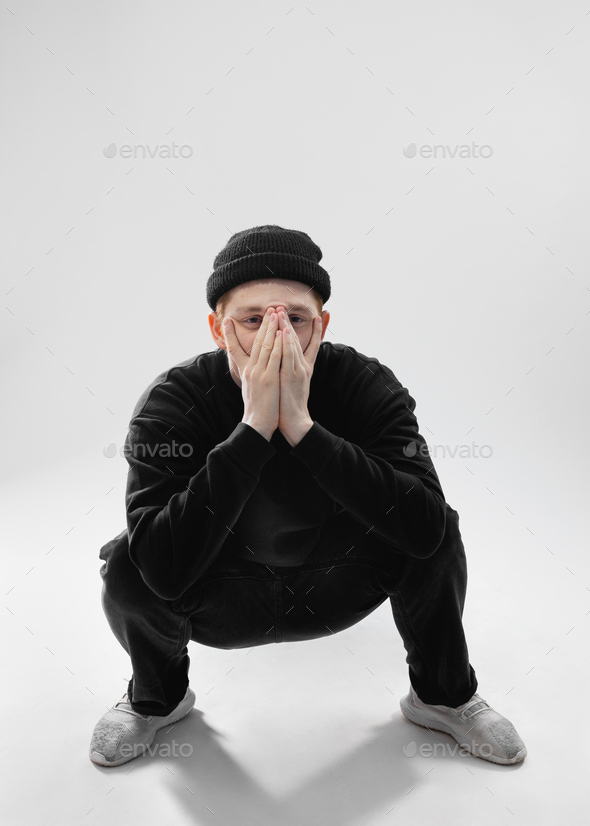 Guy dressed in black jeans, sweatshirt, hat and gray sneakers is squatting and closing his face with - Stock Photo - Images