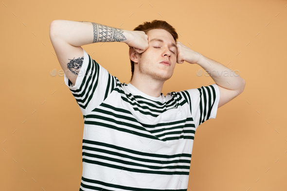Red-haired stylish guy in a striped shirt with tattoo on his hand is posing holding his hands on his
