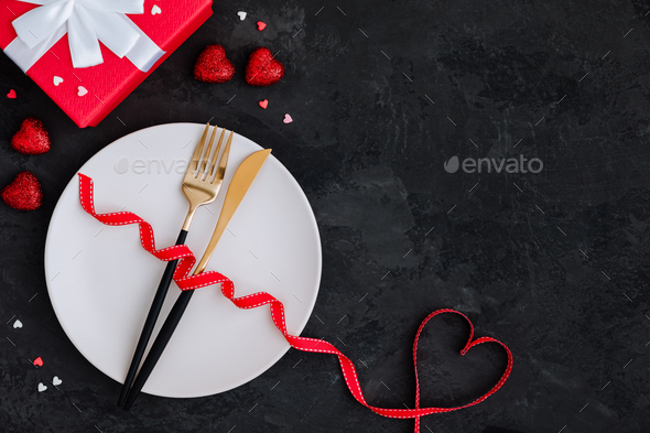 Valentine\'s Day table setting with plate, gold knife, fork, red ribbon with heart and present box