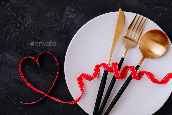 Valentine\'s Day table setting with plate, gold knife, fork, spoon, red ribbon with heart