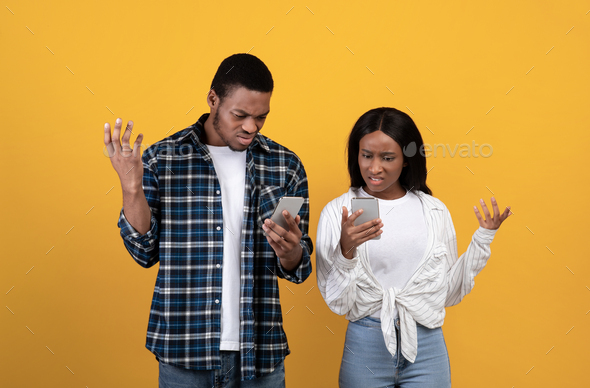 Surprised sad millennial african american couple looking at phones with open mouths and raising