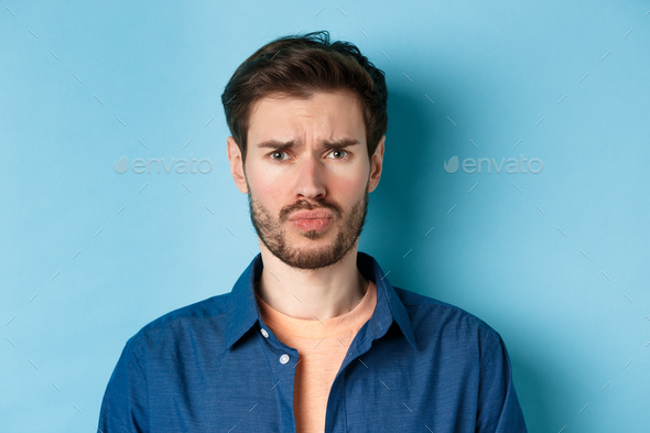 Close up of sad and offended young man sulking at someone, frowning upset, standing on blue
