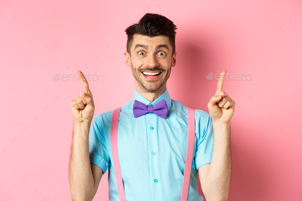Portrait of cheerful young guy with moustache and bristle pointing fingers up, showing top logo