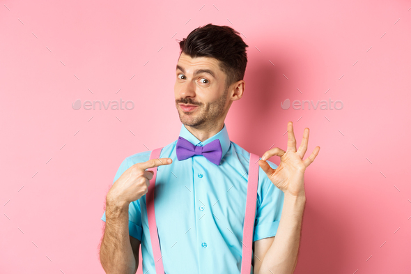Image of young handsome guy in bow-tie and holiday outfit, pointing at himself and showing okay
