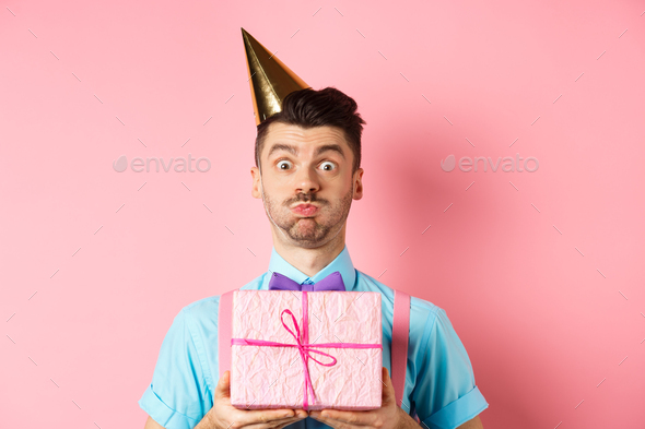 Holidays and celebration concept. Funny guy staring at camera surprised, wearing party hat, holding
