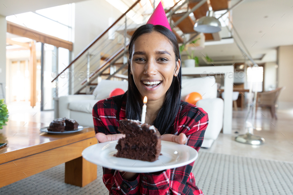 Smiling mixed race woman wearing party hat holding birthday cake having video chat