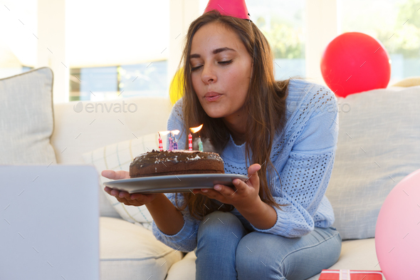 Caucasian woman having birthday video call blowing out candles on cake wearing party hat
