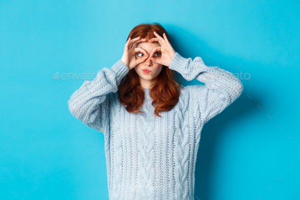 Funny redhead female model in sweater, staring at camera through fingers glasses, express interest
