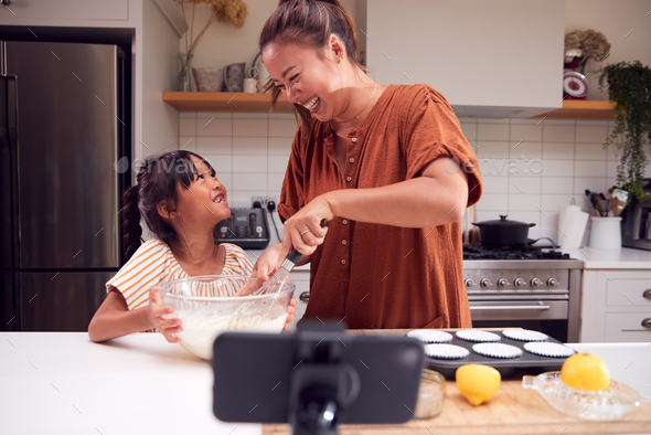 Asian Mother And Daughter Baking Cupcakes In Kitchen At Home Whilst On Vlogging On Mobile Phone - Stock Photo - Images