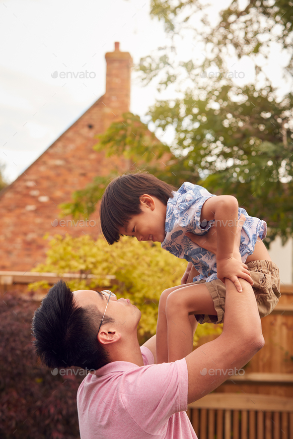 Asian Father Lifting Son In Mid Air As They Play Game In Garden Together