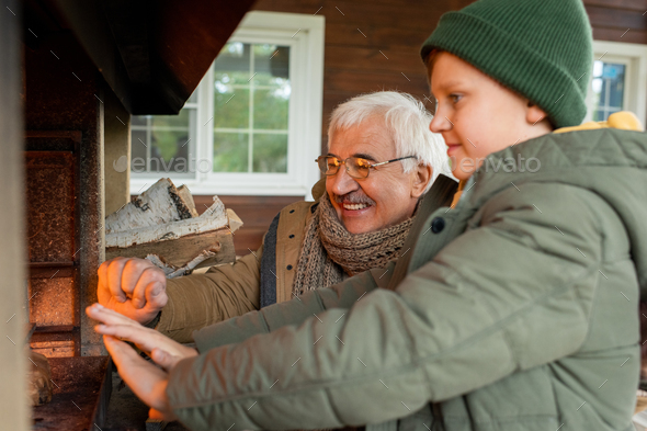 Happy senior man and his grandson standing by fireplace with burning firewoods