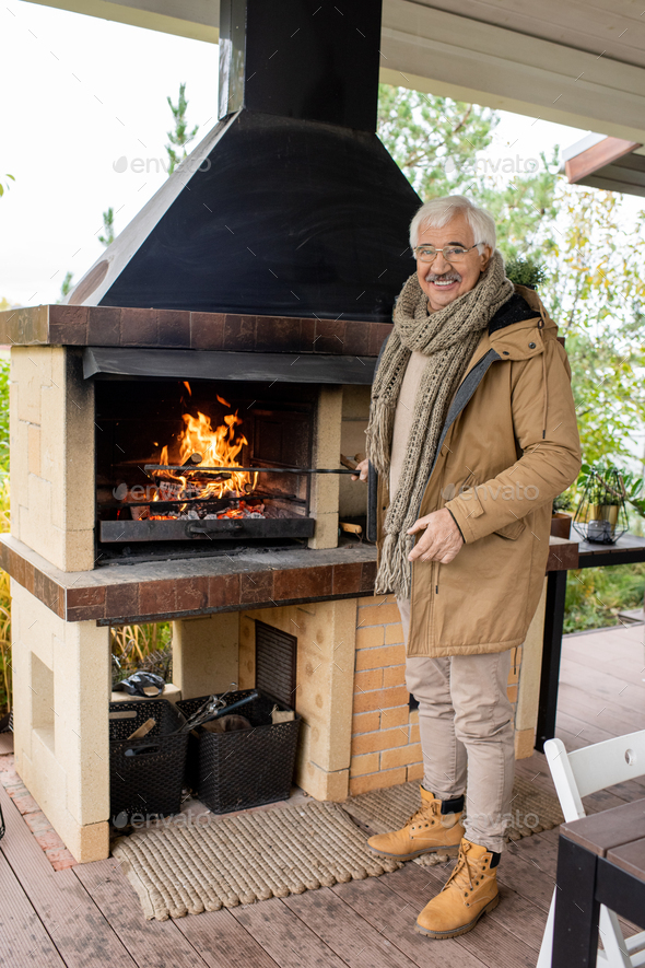 Cheerful senior man in warm casualwear standing by fireplace on patio by house