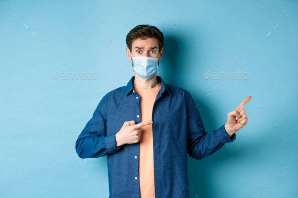 Covid and healthcare concept. Worried young man in medical mask raising eyebrow, pointing finger
