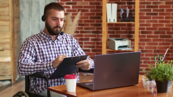 Hipster Man Wearing Headphones with Microphone is Sitting in Wheelchair Using Laptop Talking Via alt