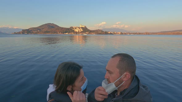 Couple in love takes off masks due to covid and kiss with lake and castle in background at sunset alt