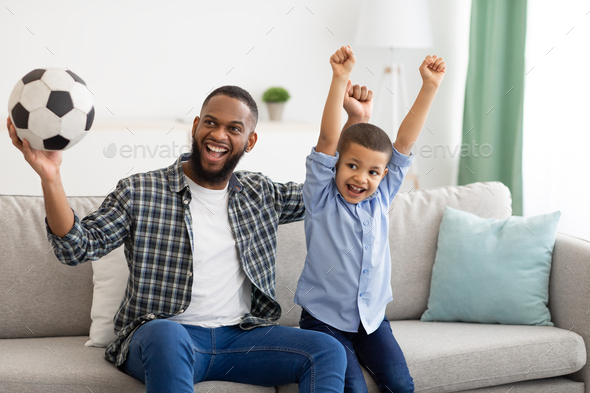 African Father And Son Watching Soccer Game On TV Indoor