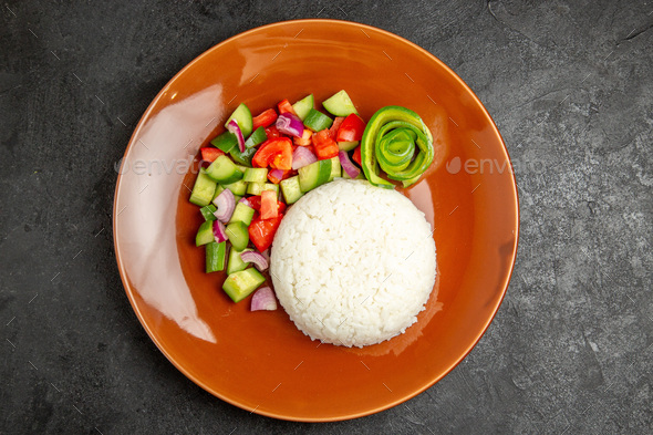 Above view of homemade rice dish and salad with tomato and cucumber on dark table