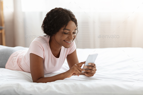 Happy black lady laying on bed, using mobile phone