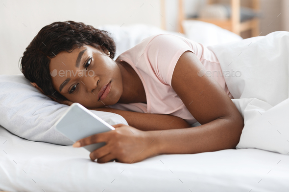 Frustrated black woman looking at smartphone while laying in bed