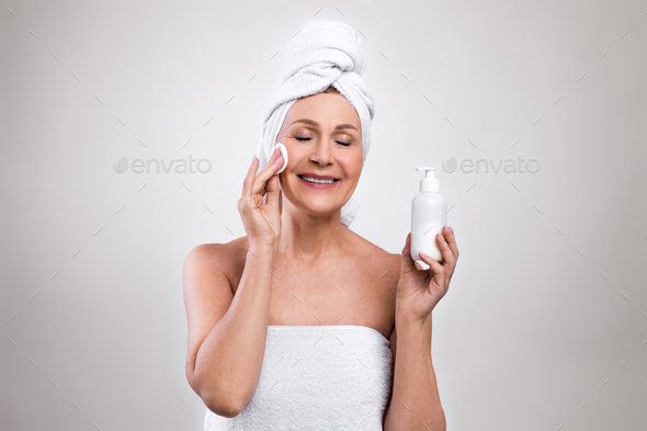 Senior woman wearing bath towels, enjoying her beauty and face care routine, applying skin tonic on