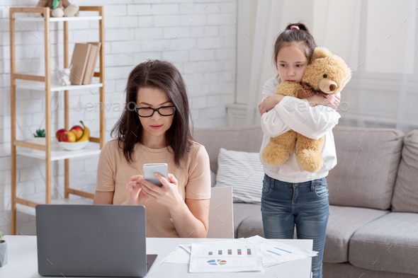 Lonely teen girl hugging teddy bear while her busy mom working online from home, not paying