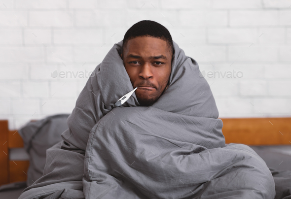 Ill African Guy Holding Thermometer In Mouth Sitting In Bedroom