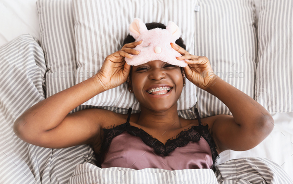 Lazy cheerful millennial african american female takes off sleep mask and looks at camera, lies on