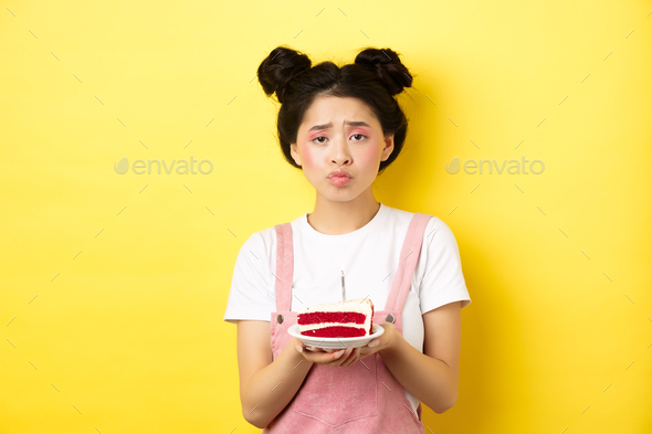 Sad and lonely birthday girl frowning upset, holding birthday cake with candle, making wish