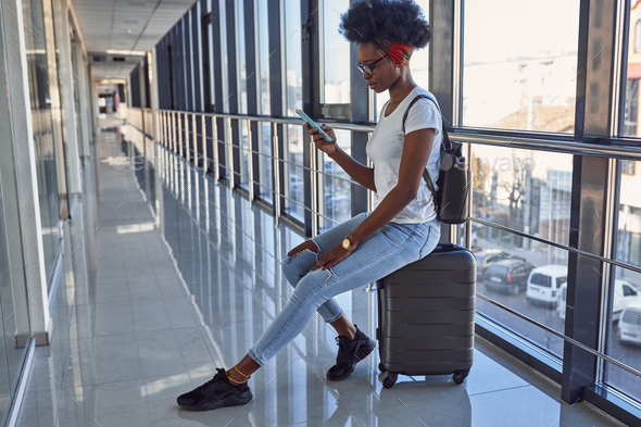 Young african american female passanger in casual clothes is in airport with baggage. Using phone