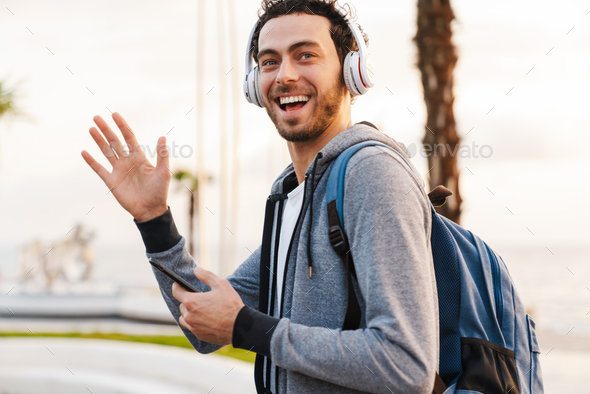 Smiling guy in headphones waving hand and using mobile phone