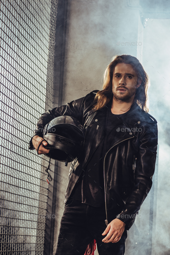 Stylish long haired bearded man in leather jacket holding helmet