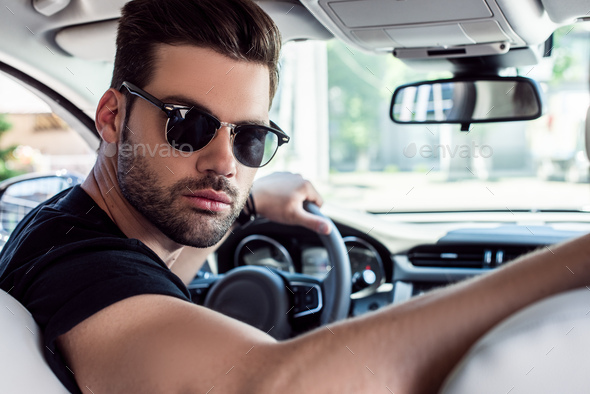 portrait of serious stylish man in sunglasses looking back while driving  car Stock Photo by LightFieldStudios
