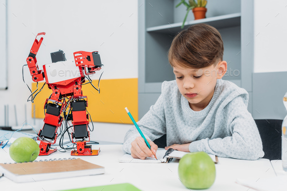 focused schoolboy sitting at desk with robot model and writing in notebook during STEM lesson