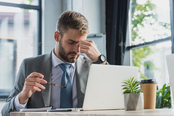 tired young businessman holding eyeglasses and rubbing nose bridge while using laptop in office
