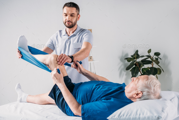 rehabilitation therapist assisting senior man exercising with rubber tape on massage table