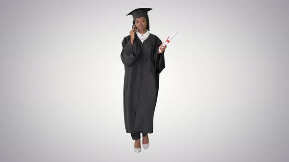 African American Female Student in Graduation Robe Talking on the Phone While Walking on Gradient alt