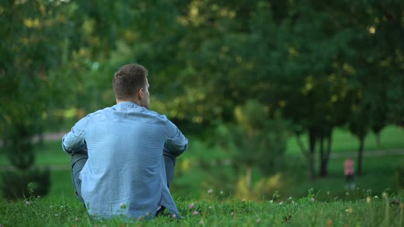 Back View of Man Sitting on Grass at Park, Enjoying Weekend in Loneliness alt