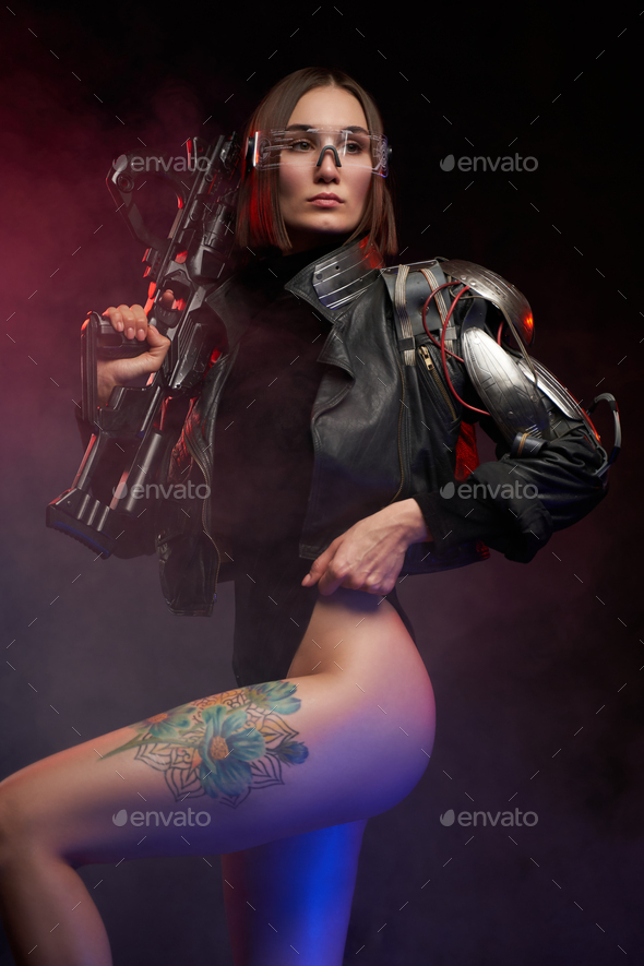 Sexy and slim woman with implant and rifle poses in dark background