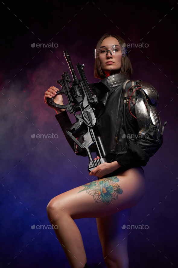 Woman with cybernetic arm poses with rifle in dark background