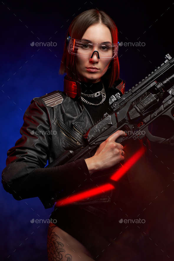 Fashionable woman with stylish haircut in black clothing poses holding a  futuristic rifle. Beautiful female soldier in cyberpunk style. Stock Photo