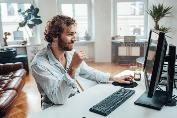 Joyful guy with curly hairs and beard sits at table working on pc