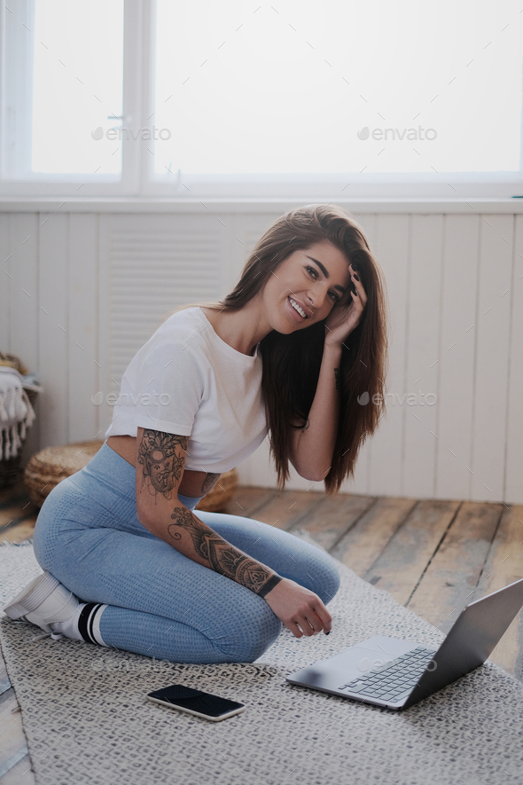 Positive and tattooed woman with laptop in room with sunbeams