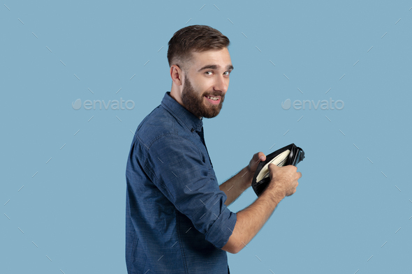 Funky Caucasian guy holding full wallet with money on blue studio background