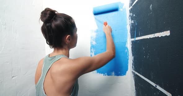 Rear View of Attractive Young Woman Painting White Wall Onto Blue with Roller alt