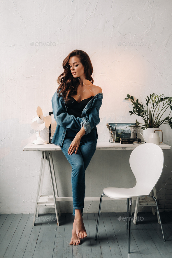 a brunette with long hair poses in the interior sitting on a table Stock  Photo by Lobachad